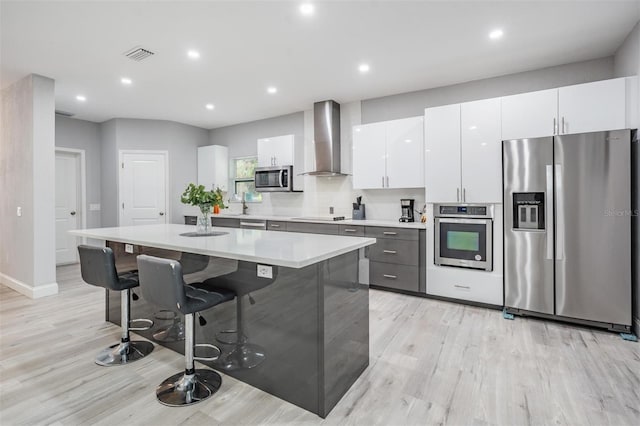 kitchen with appliances with stainless steel finishes, a kitchen island, light hardwood / wood-style floors, wall chimney range hood, and white cabinetry