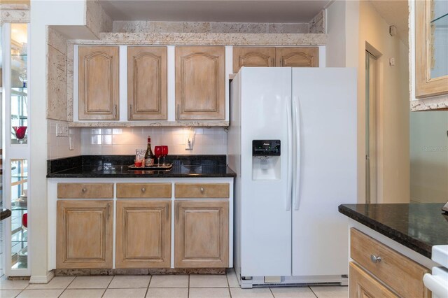 kitchen with light tile patterned floors, white refrigerator with ice dispenser, dark stone countertops, and backsplash