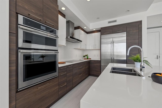 kitchen with appliances with stainless steel finishes, white cabinetry, wall chimney exhaust hood, backsplash, and sink