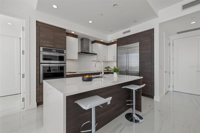 kitchen featuring light tile floors, appliances with stainless steel finishes, a breakfast bar, an island with sink, and wall chimney range hood