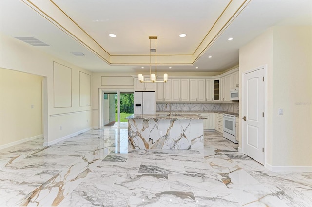 kitchen featuring light stone countertops, a raised ceiling, white appliances, hanging light fixtures, and a center island with sink