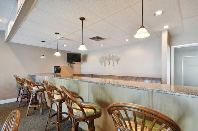 kitchen featuring a drop ceiling, light stone counters, dark carpet, hanging light fixtures, and a kitchen breakfast bar