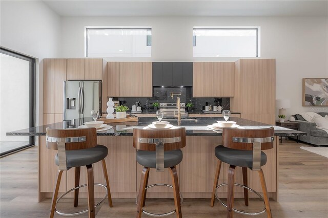 kitchen featuring a kitchen island with sink, dark stone countertops, stainless steel refrigerator with ice dispenser, and a breakfast bar area