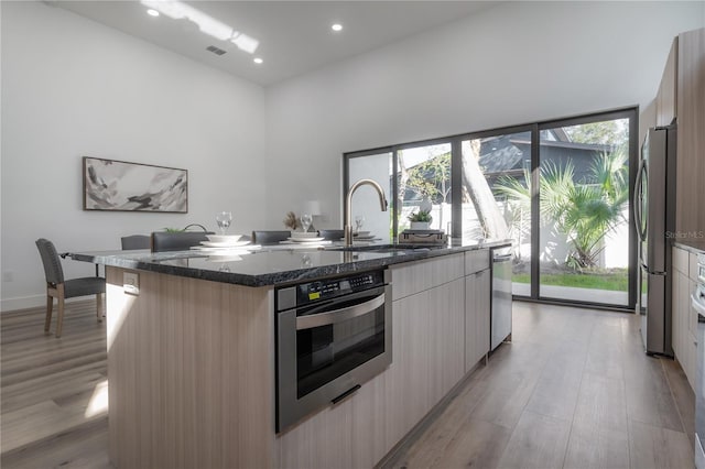 kitchen with sink, light hardwood / wood-style flooring, dark stone countertops, an island with sink, and appliances with stainless steel finishes