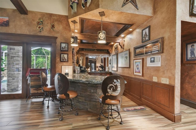 bar with beam ceiling, a wainscoted wall, hanging light fixtures, and light wood-style flooring