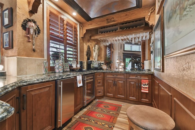 kitchen featuring light wood finished floors, beverage cooler, wainscoting, brown cabinets, and dark stone countertops