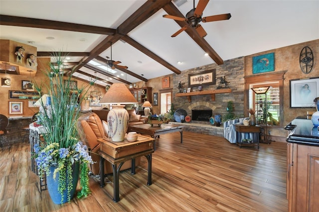 living room with lofted ceiling with beams, a stone fireplace, wood finished floors, and a ceiling fan