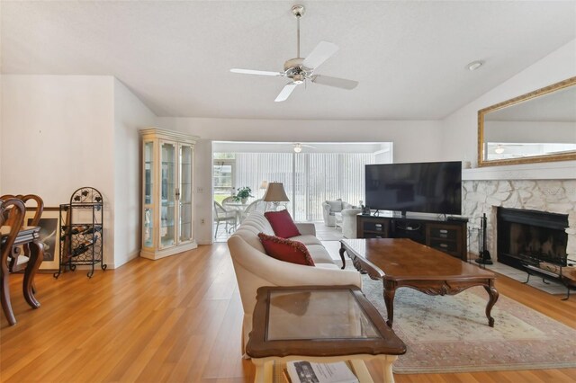 living room with ceiling fan, lofted ceiling, a stone fireplace, and light wood-type flooring