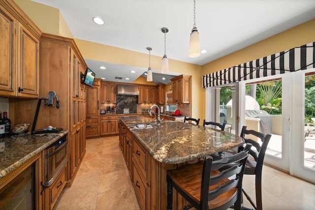 kitchen featuring tasteful backsplash, an island with sink, light tile flooring, dark stone countertops, and wall chimney exhaust hood