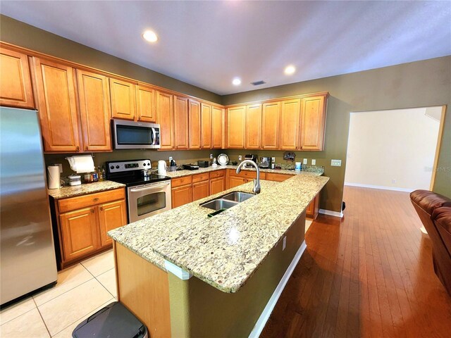 kitchen featuring a kitchen island with sink, sink, light stone countertops, appliances with stainless steel finishes, and light hardwood / wood-style floors