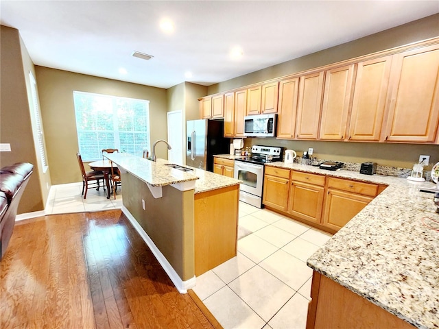 kitchen featuring appliances with stainless steel finishes, an island with sink, sink, light stone counters, and light hardwood / wood-style flooring