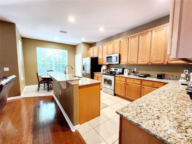 kitchen with a kitchen island with sink, sink, light hardwood / wood-style flooring, light stone countertops, and appliances with stainless steel finishes