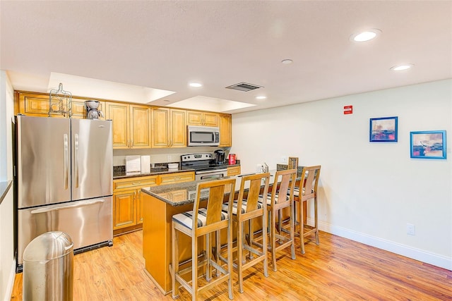 kitchen with appliances with stainless steel finishes, light wood-type flooring, dark stone counters, a raised ceiling, and a breakfast bar area