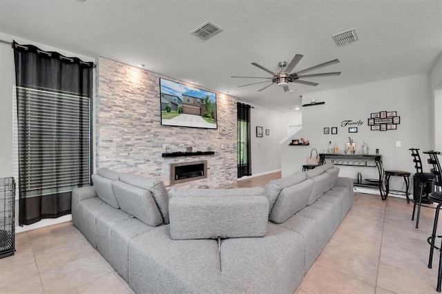 living room with ceiling fan, a stone fireplace, light tile patterned floors, and a textured ceiling