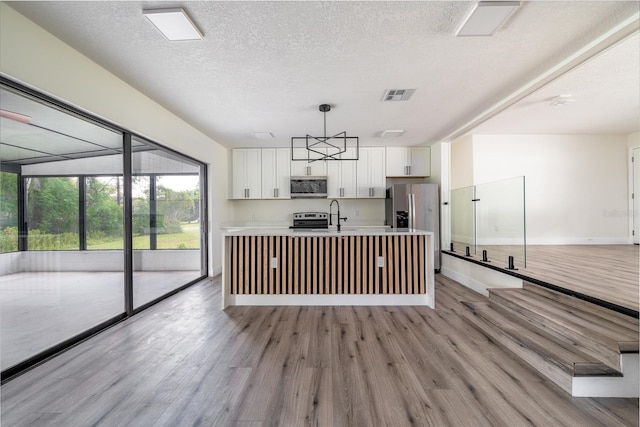 kitchen with decorative light fixtures, white cabinetry, stainless steel appliances, a center island with sink, and light wood-type flooring