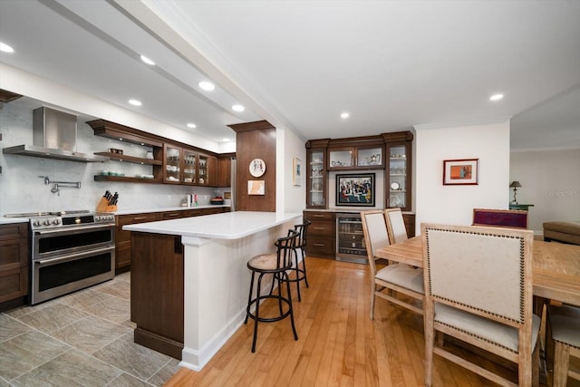 kitchen featuring a breakfast bar area, dark brown cabinetry, light countertops, wall chimney range hood, and double oven range