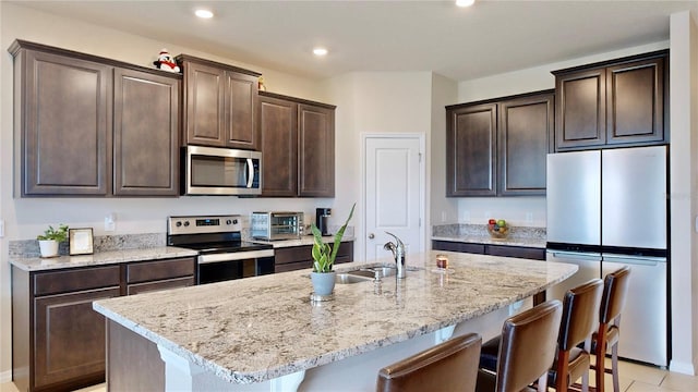 kitchen featuring dark brown cabinetry, a center island with sink, light stone counters, and appliances with stainless steel finishes