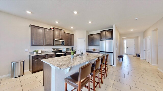 kitchen with light stone countertops, dark brown cabinetry, a breakfast bar, a kitchen island with sink, and appliances with stainless steel finishes