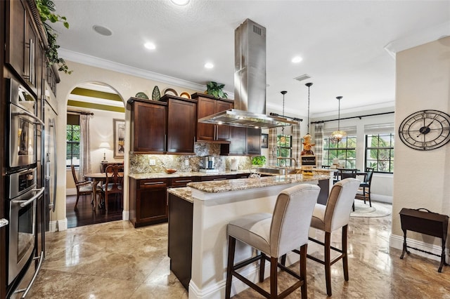kitchen featuring crown molding, island range hood, decorative backsplash, a breakfast bar, and arched walkways