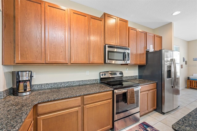 kitchen with light tile patterned floors, appliances with stainless steel finishes, brown cabinetry, a textured ceiling, and dark stone countertops