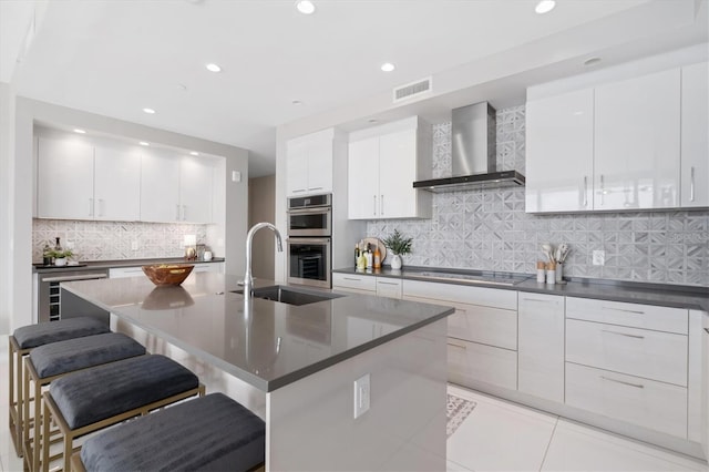 kitchen with white cabinetry, a kitchen island with sink, double oven, and wall chimney range hood