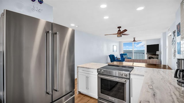 kitchen with white cabinetry, light hardwood / wood-style flooring, premium appliances, a stone fireplace, and ceiling fan with notable chandelier