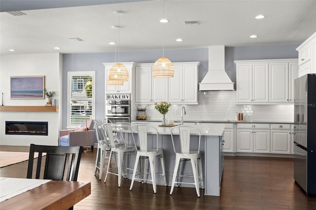 kitchen with a kitchen island with sink, premium range hood, hanging light fixtures, appliances with stainless steel finishes, and white cabinetry