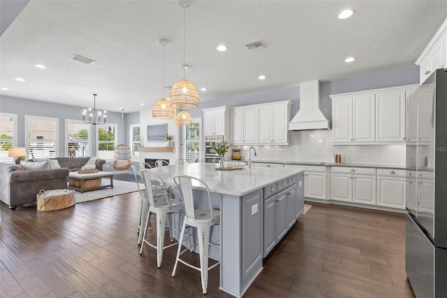 kitchen featuring backsplash, custom exhaust hood, pendant lighting, white cabinetry, and an island with sink