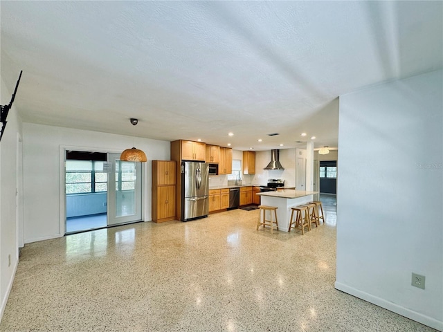 kitchen featuring a center island, wall chimney range hood, hanging light fixtures, appliances with stainless steel finishes, and a kitchen bar