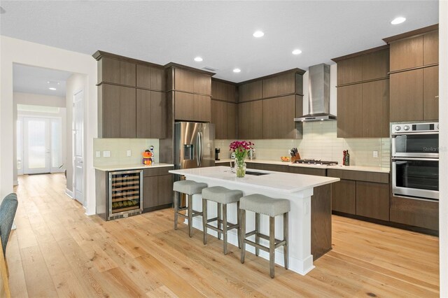 kitchen featuring wine cooler, a kitchen breakfast bar, stainless steel appliances, wall chimney range hood, and light hardwood / wood-style flooring