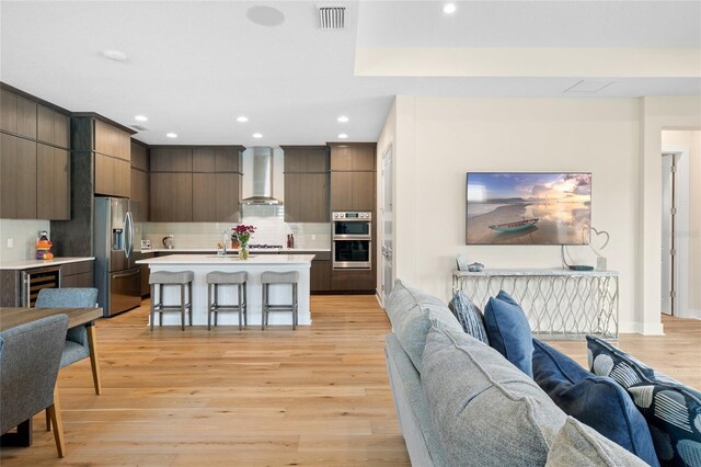 interior space with appliances with stainless steel finishes, an island with sink, a kitchen bar, wall chimney exhaust hood, and light wood-type flooring