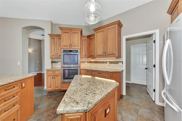 kitchen featuring light stone countertops, stainless steel double oven, white fridge, a kitchen island, and hanging light fixtures