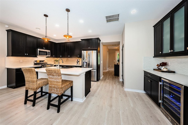 kitchen featuring stainless steel appliances, beverage cooler, a kitchen island with sink, pendant lighting, and light hardwood / wood-style flooring