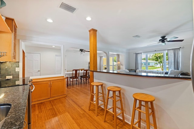 kitchen featuring ornate columns, light hardwood / wood-style flooring, dark stone countertops, decorative backsplash, and a breakfast bar