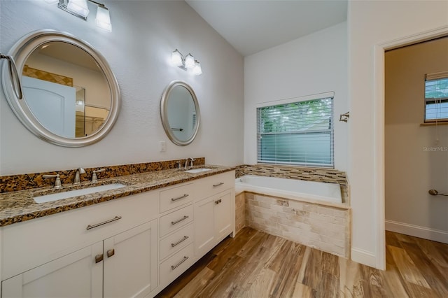 bathroom with dual vanity, tiled bath, and wood-type flooring