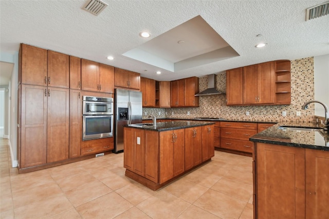 kitchen with sink, appliances with stainless steel finishes, an island with sink, dark stone counters, and wall chimney range hood