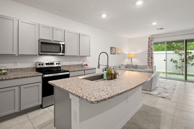 kitchen with sink, light stone counters, gray cabinets, an island with sink, and stainless steel appliances