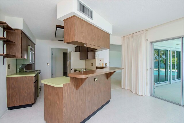 kitchen featuring white appliances, light tile patterned floors, kitchen peninsula, backsplash, and a kitchen bar