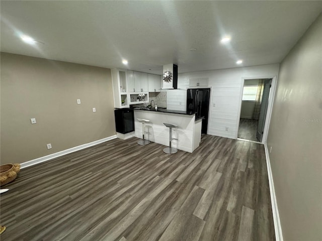 kitchen featuring wall chimney exhaust hood, a kitchen bar, black refrigerator, white cabinetry, and kitchen peninsula