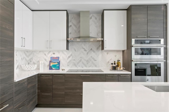 kitchen with white cabinetry, stainless steel double oven, black electric stovetop, decorative backsplash, and wall chimney range hood