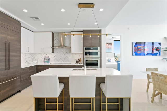 kitchen with a kitchen bar, hanging light fixtures, a center island with sink, stainless steel double oven, and wall chimney range hood