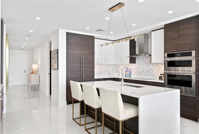 kitchen featuring a breakfast bar, decorative light fixtures, a center island with sink, double oven, and wall chimney range hood