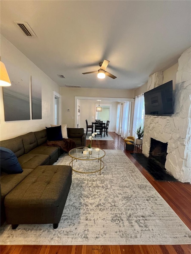 living room featuring dark wood-type flooring, ceiling fan, and a stone fireplace