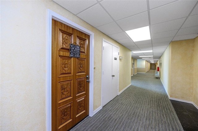 corridor featuring a paneled ceiling, dark carpet, and baseboards