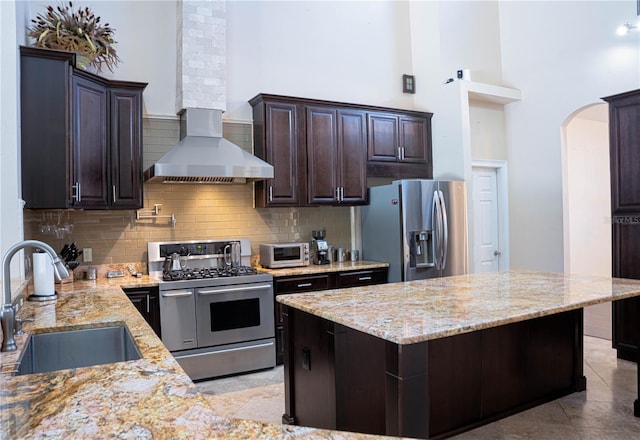 kitchen featuring wall chimney exhaust hood, dark brown cabinetry, sink, light stone counters, and appliances with stainless steel finishes