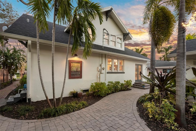 front of house at dusk with roof with shingles, fence, and stucco siding