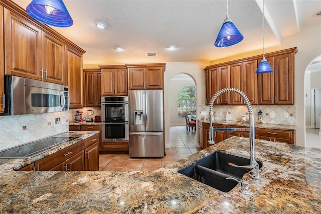 kitchen featuring arched walkways, stainless steel appliances, brown cabinetry, a sink, and dark stone countertops