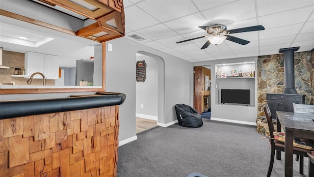 living room with ceiling fan, a drop ceiling, dark carpet, and a wood stove