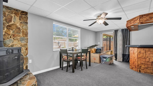 dining space with ceiling fan, a drop ceiling, carpet floors, and a wood stove