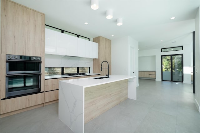 kitchen with white cabinetry, sink, light stone counters, an island with sink, and double oven